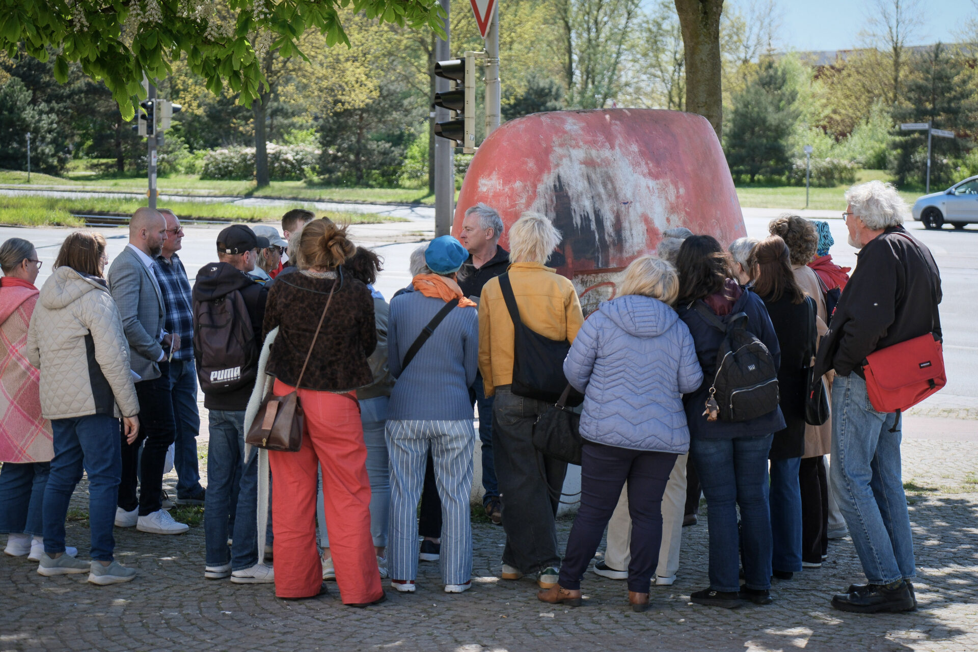 Auf einem öffentlichen Platz haben sich rund um einen Sprecher und eine Skulptur Menschen versammelt, die zuhören.