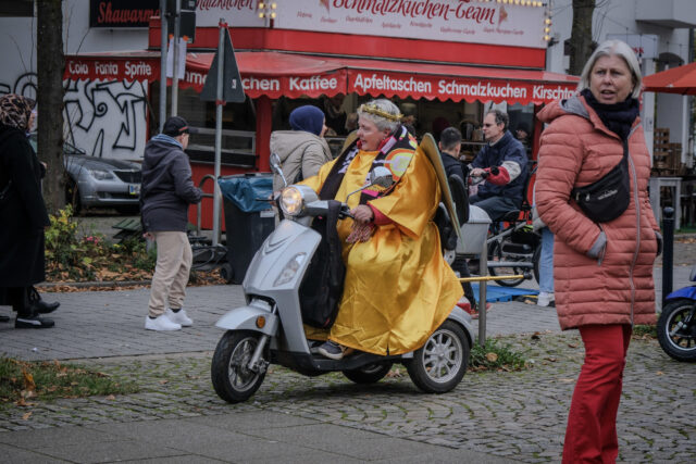 Auf einer belebten Straße fährt eine Frau auf einem Moped. Die Frau ist verkleidet als Engel, sie trägt einen goldenen Mantel, eine goldene Krone und auf ihrem Rücken große Flügel. IM Hintergrund sind mehrere vorbeilaufende Menschen zu sehen und ein Straßenstand auf dem "Kaffee, Apfeltaschen, Schmalzkuchen" steht.