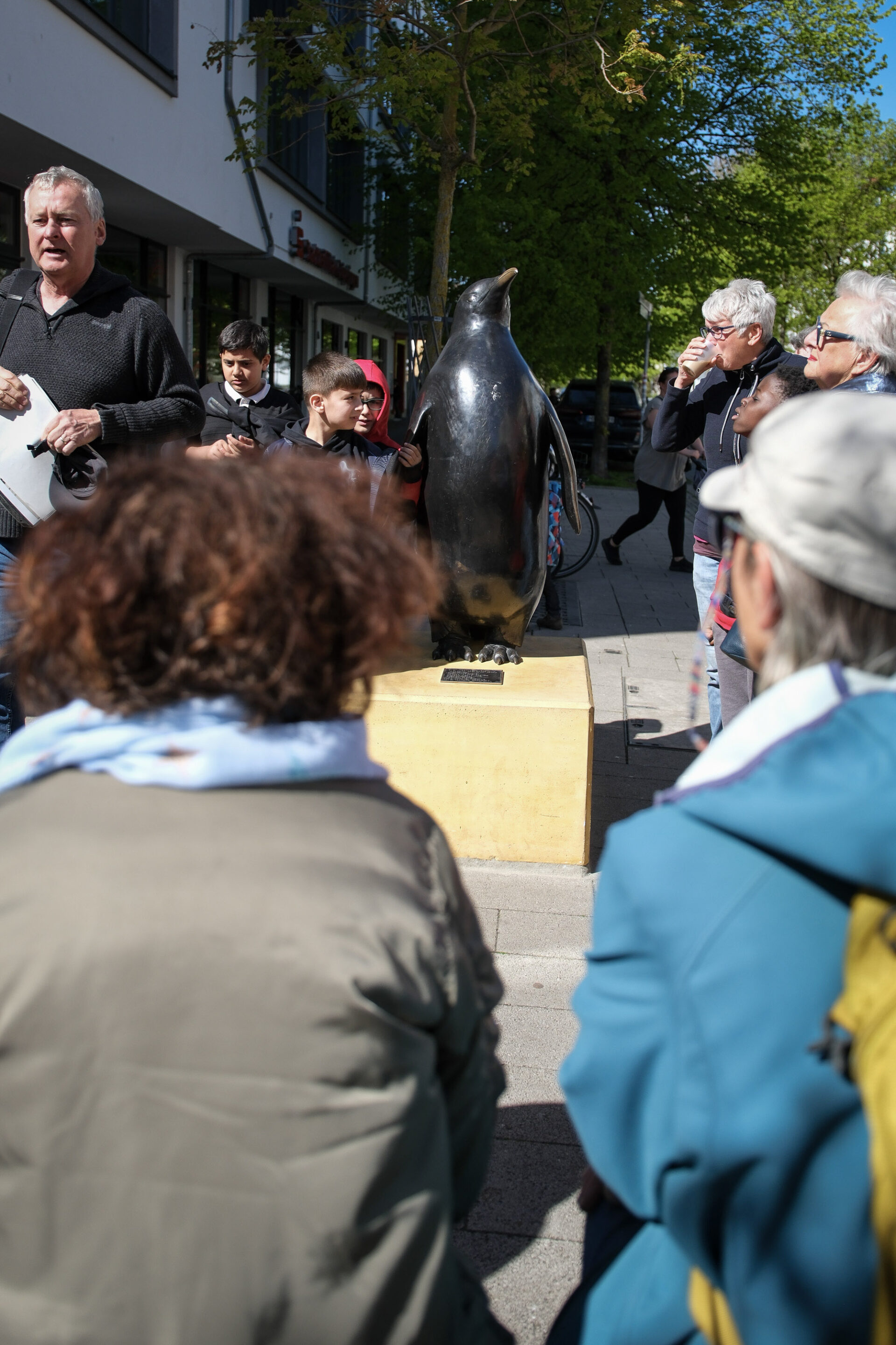 Menschen sitzen und stehen rund um einen Redner und eine Skulptur.