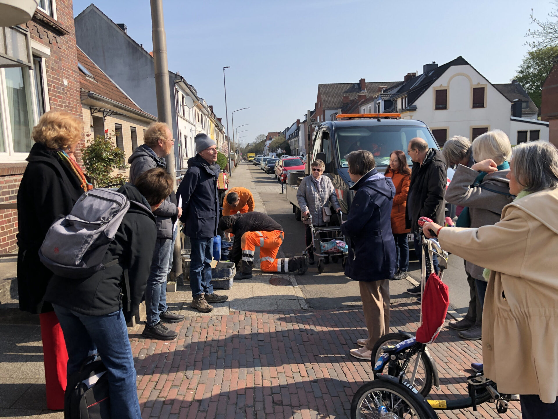 Eine Menschentrabe steht in einer Straße, bei der Handwerker einen Stolperstein verlegen. Eine Person spricht. 