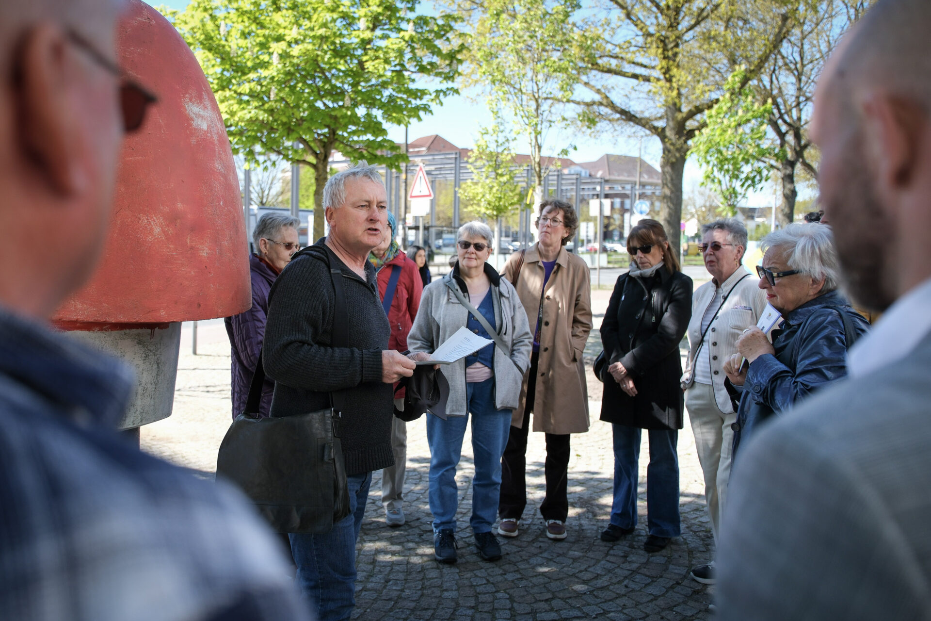 Auf einem öffentlichen Platz haben sich rund um einen Sprecher und eine Skulptur Menschen versammelt, die zuhören.