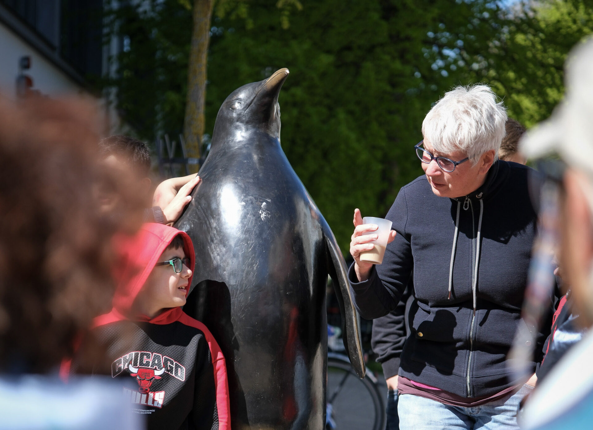 Rund um eine Bronzeskulptur in Form eines Pinguins steht eine Personengruppe. Auf dem Sockel des Pinguins sitzt ein Kind im roten Sweater.