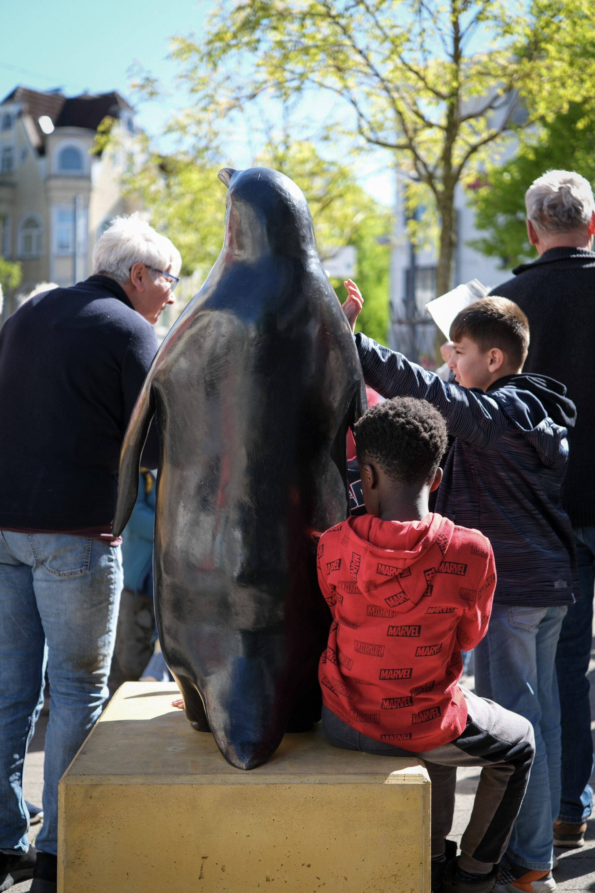 Rund um eine Bronzeskulptur in Form eines Pinguins steht eine Personengruppe. Auf dem Sockel des Pinguins sitzt ein Kind im roten Sweater.