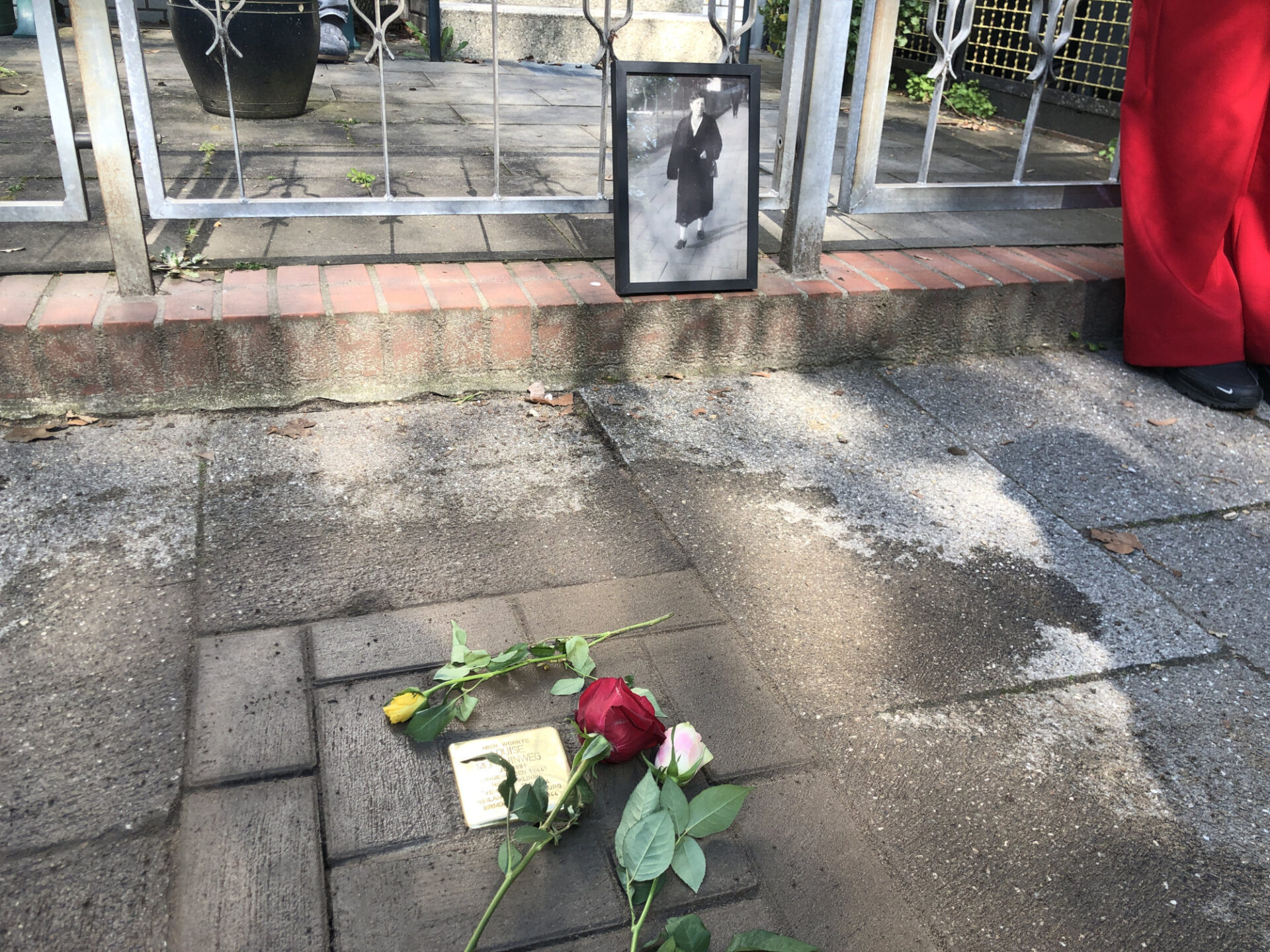 Ein Stolperstein, umrahmt von Blumen. Ein gerahmtes Bild erinnert an die Person. 