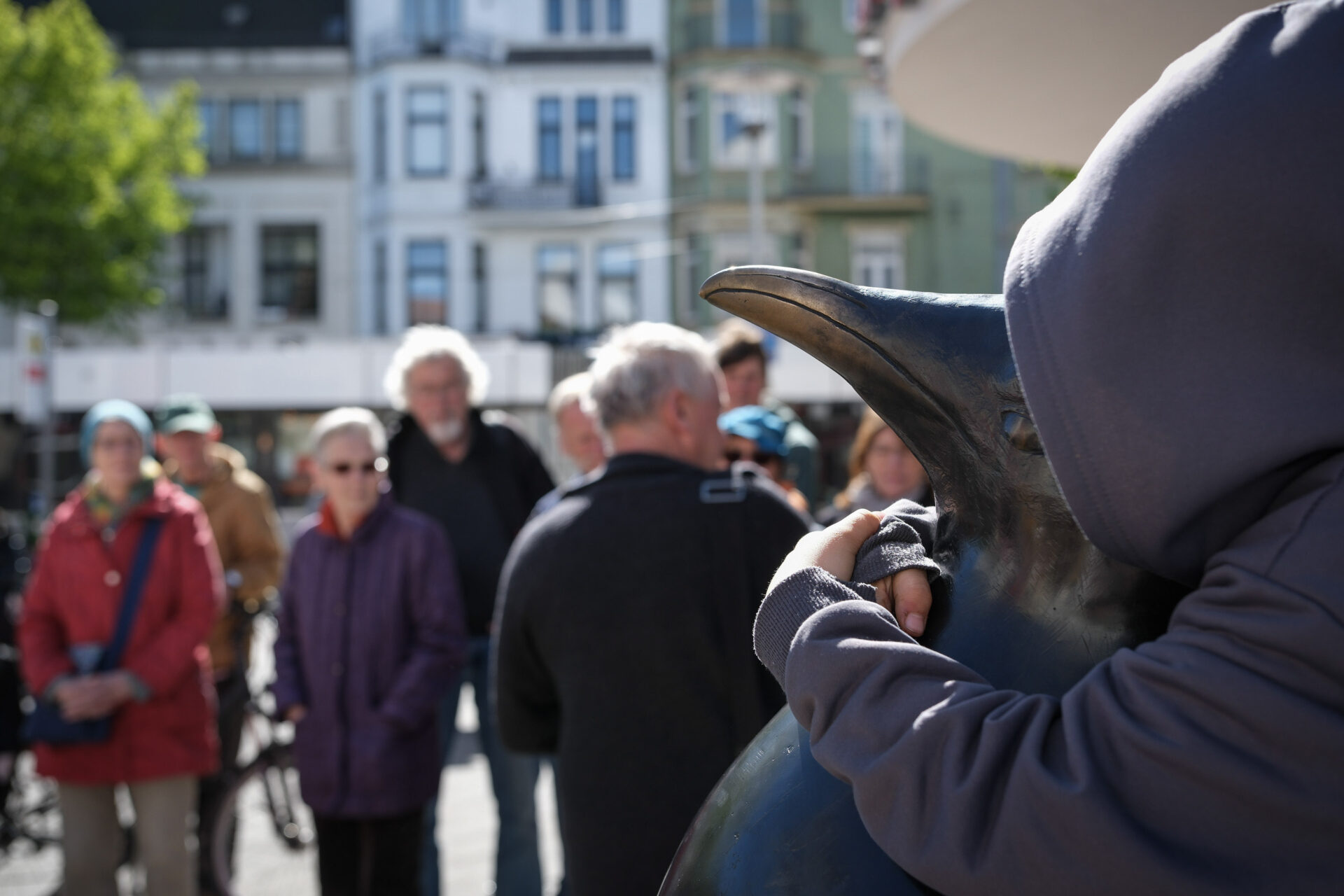 Ein Kind im Hoody umarmt den Hals einer Pinguin-Bronzeskulptur. Hintergrund sind Menschen.