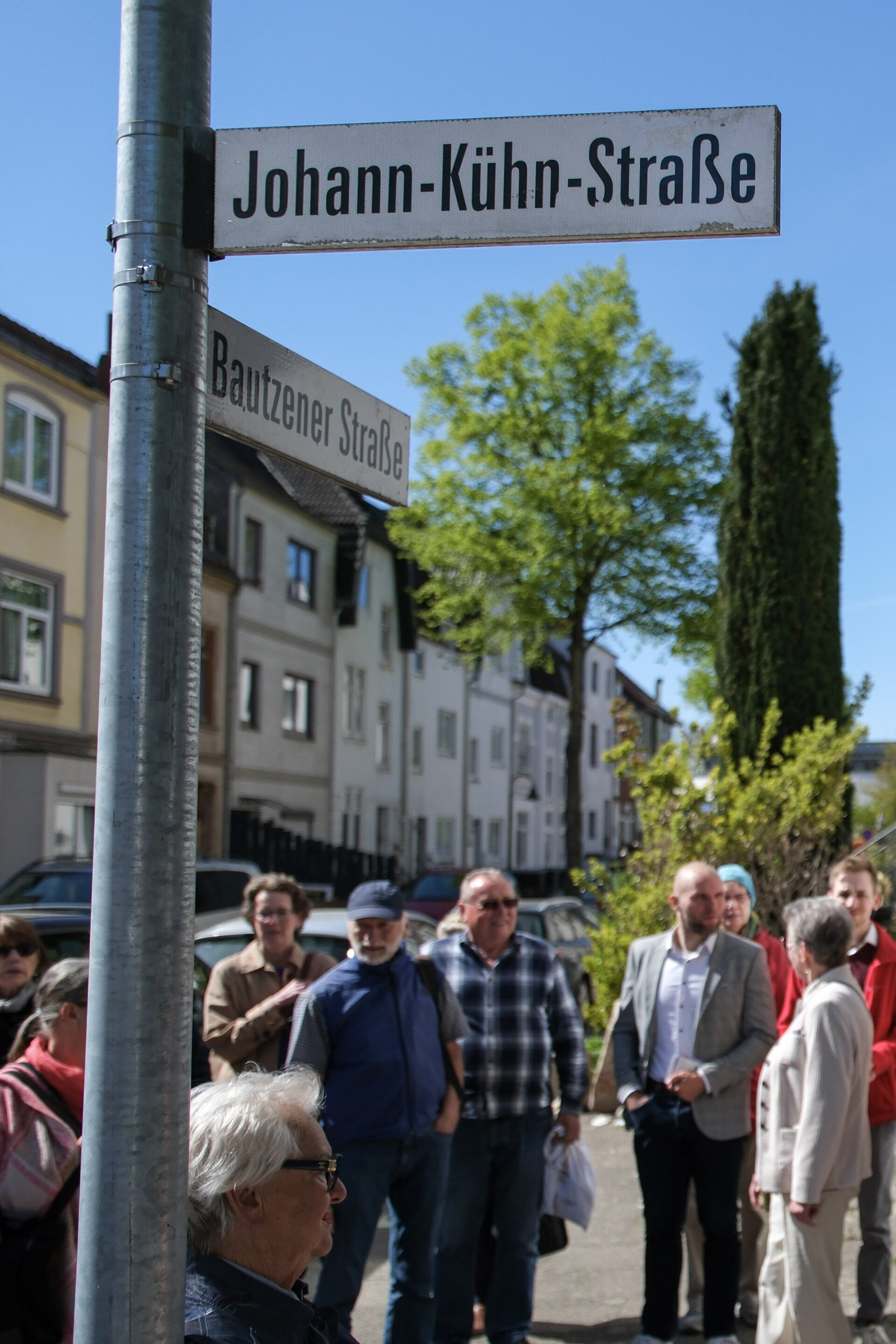 Im Vordergrund ist das Straßenschild "Johann-Kühn-Straße" zu sehen. Im Hintergrund befindet sich eine Menschenmenge.