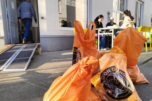 Orangefarbene Müllsäcke mit schwarzem Logo liegen auf einem Haufen. Im Hintergrund ist der Eingang zu einem Gebäude zu sehen. Eine mobile Rampe ist über die Stufen im Eingangsbereich gelegt. Rechts sitzen Personen um einen Tisch im Außenbereich.
