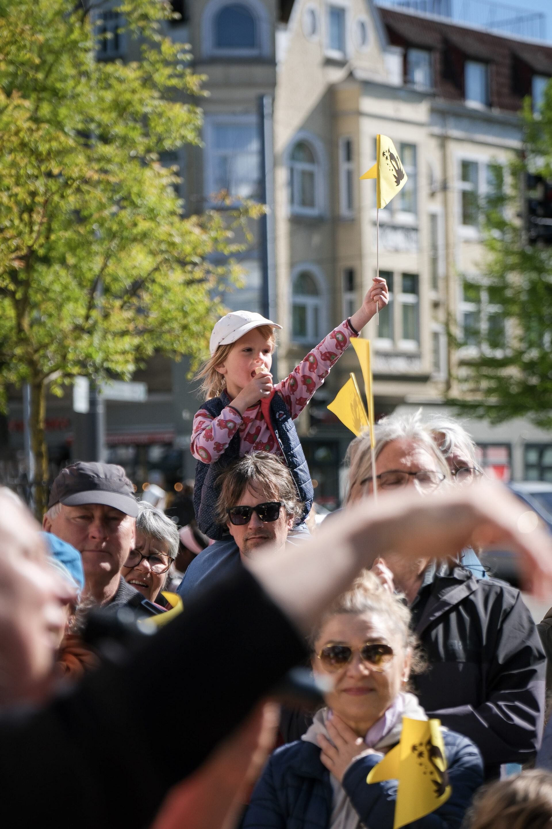 Ein Kind sitzt auf den Schultern eines Mannes und hält in der einen Hand ein gelbes Fähnchen hoch und in der anderen ein Eis.