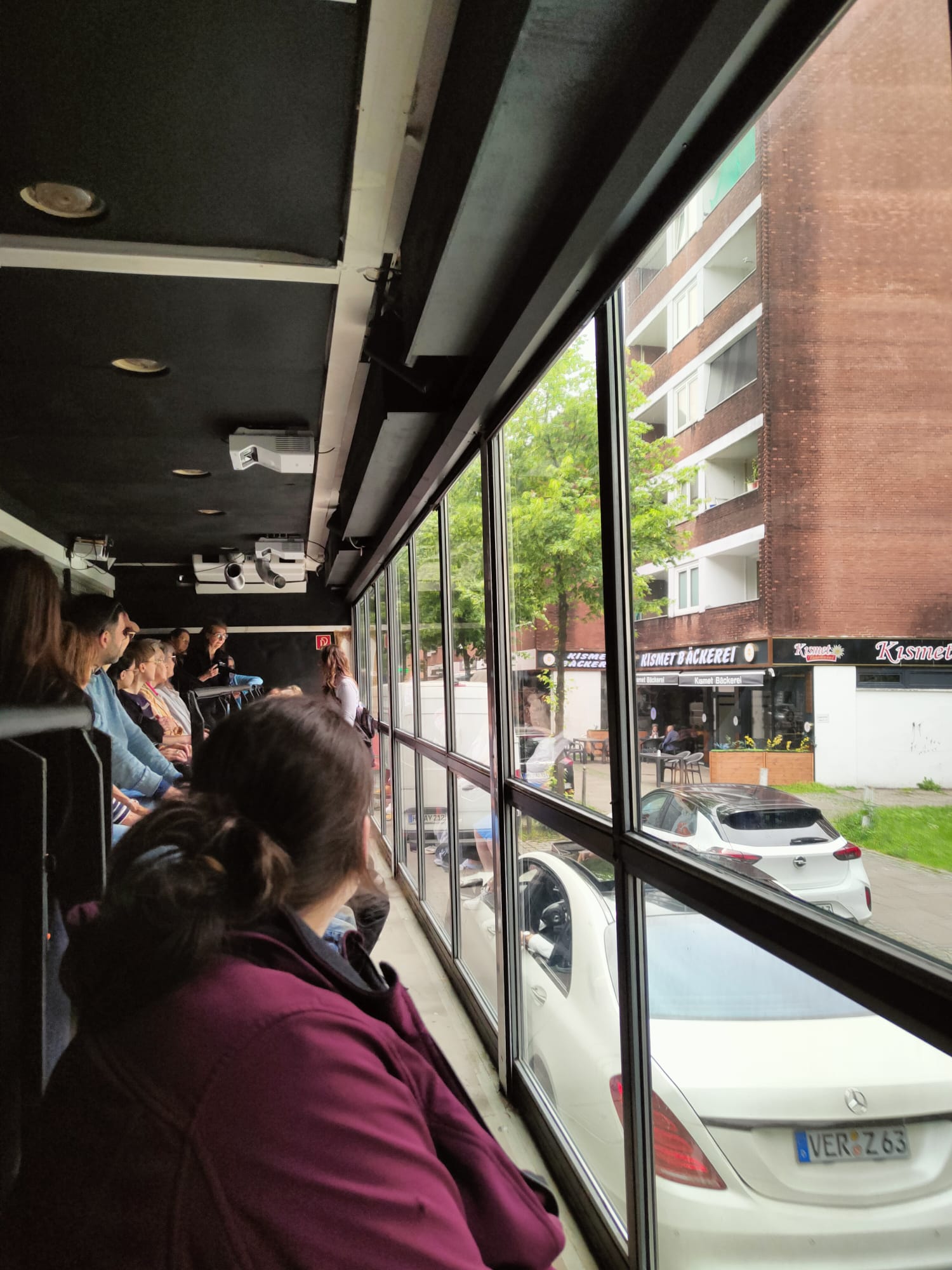 Menschen sitzen in Reihen in einem gläsernen Bus und schauen raus auf die Straße durch rieseige Glaswände