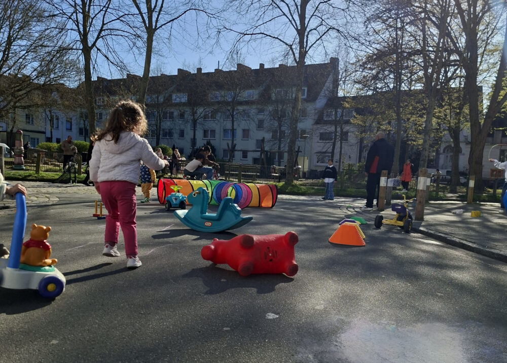 Straßenspielzeug liegt verteilt auch deiner Straßenkreuzung. Ein Kind ist von hinten zu sehen. Im Hintergrund befinden sich eine Person, Bäume und Häuser.