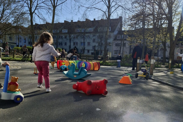 Straßenspielzeug liegt verteilt auch deiner Straßenkreuzung. Ein Kind ist von hinten zu sehen. Im Hintergrund befinden sich eine Person, Bäume und Häuser.
