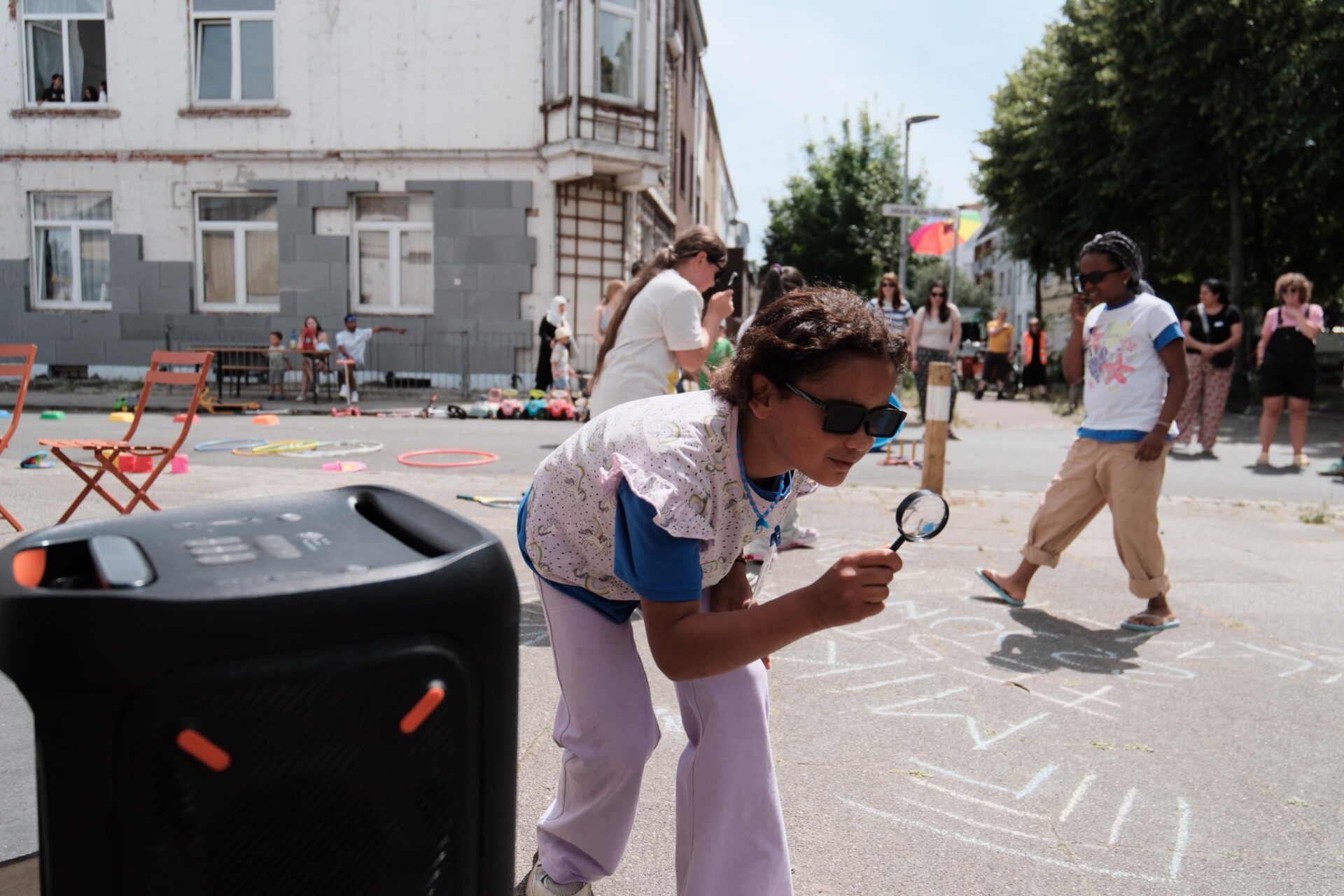 Ein Kind mit Sonnenbrille sucht auf der Straße mit einer Lupe nach vorn gebeugt nach etwas. Im Hintergrund sind noch mehr Kinder zu sehen, die spielen.