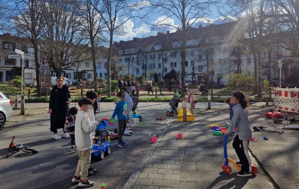 Kinder spielen auf der Straße im Rahmen einer Spielstraßenaktion. Im hintergrund sind Bäume und Häuser zu sehen.