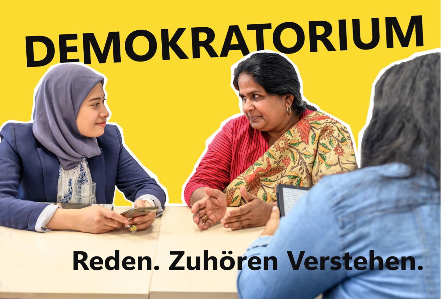 Drei Personen sitzen an einem Tisch. Links sitzt eine Person mit grauem Kopftuch und dunklem Blazer und hält ein Smartphone in der Hand. In der Mitte sitzt eine Person mit rotem Oberteil und gemustertem Tuch über der Schulter, die ihre Hände vor sich auf dem Tisch bewegt. Rechts ist eine Person von hinten zu sehen, die ein Tablet oder Smartphone hält. Der Hintergrund ist gelb. Oben steht der Schriftzug „DEMOKRATORIUM“, unten der Text „Reden. Zuhören. Verstehen.“