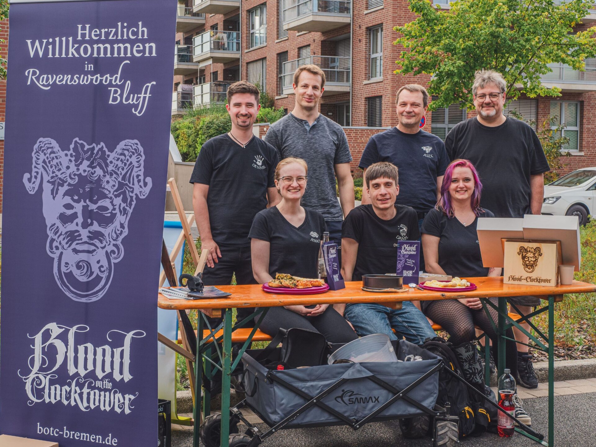 DIe Spielgruppe von "Blood on the Clocktower" sitzt am Tisch und lacht in die Kamera. Vor dem Tisch haben sie einen großen Aufsteller mit dem Symbol und der Aufschrift ihres Spiels.