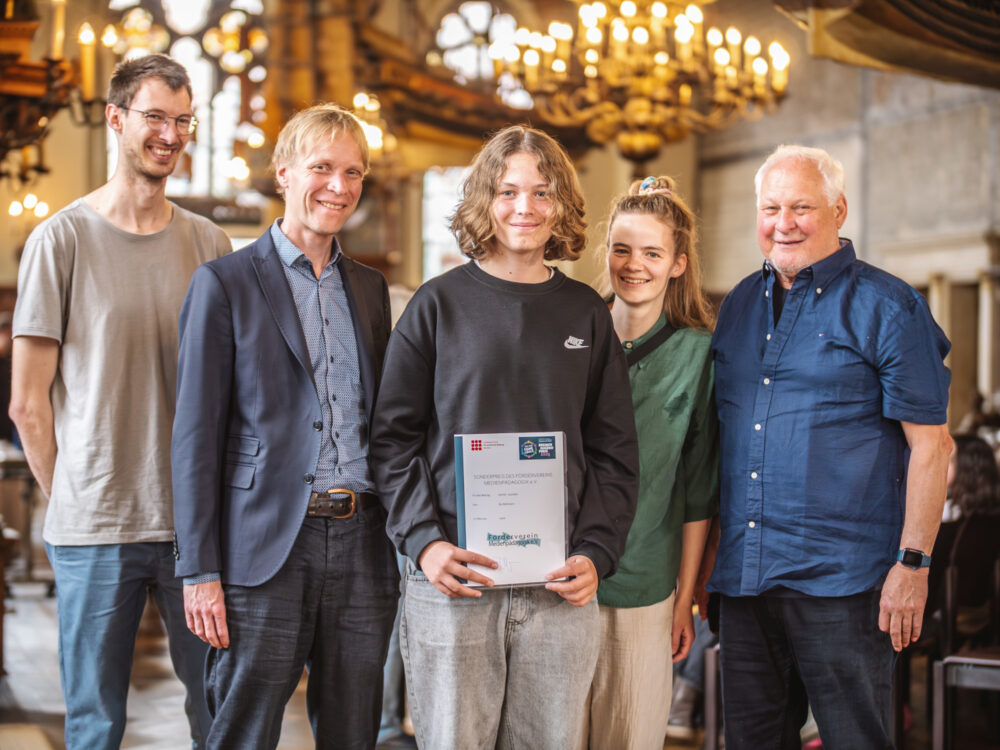 Eine Familie steht zu fünft zusammen und lächelt in die Kamera. Die junge Person in ihrer Mitte hält eine Urkunde in den Händen. Im Hintergrund ist ein Kronleuchter zu sehen.