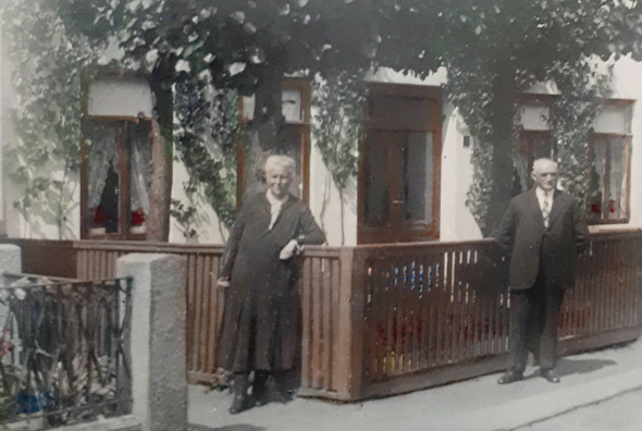 Eine ältere Frau und ein älterer Mann stehen vor einem Haus mit einer hellen Fassade und mehreren Fenstern, die von Kletterpflanzen umrahmt sind. Beide stehen hinter einem niedrigen Holzzaun auf dem Gehweg. Die Frau trägt ein dunkles Kleid, der Mann einen dunklen Anzug.