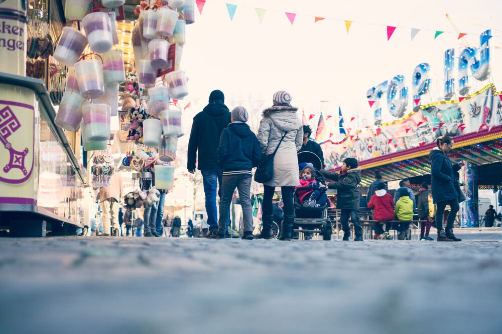 Mehrere Personen sind von hinten zu sehen. sie sind dick angezogen und haben Mützen auf dem Kopf. Links und rechts von Ihnen befinden sich Kirmesstände mit Zuckerwatte und Spielzeug.