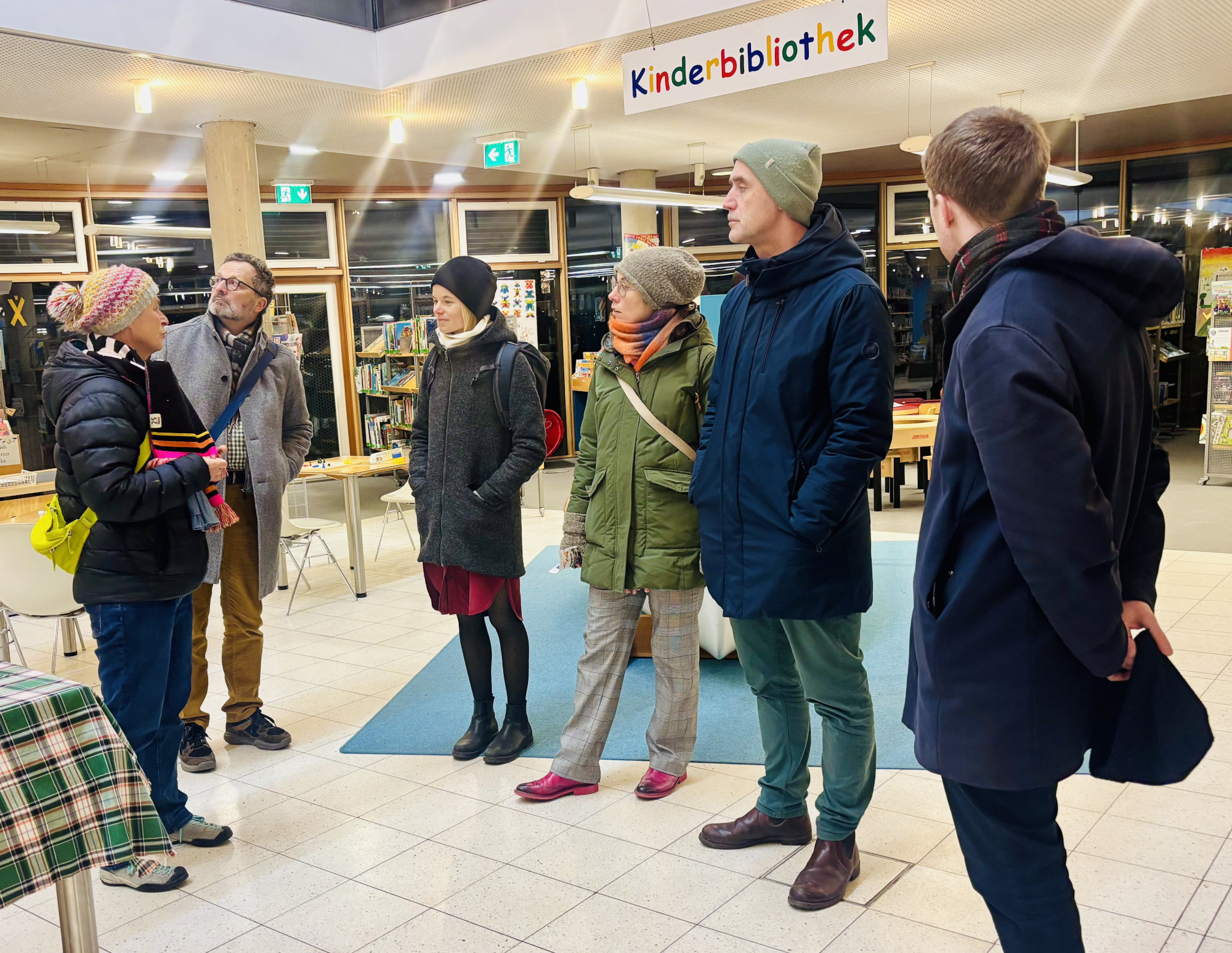 Sechs warm angezogene Personen befinden sich in einem Raum und stehen im Halbkreis. Eine Person spricht, die anderen hören zu und schauen sich um.