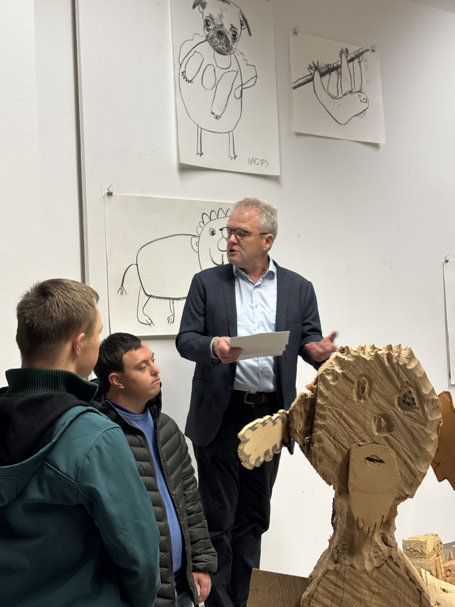 Eine Person steht vor einer Wand auf der Zeichnungen angebracht sind und hält ein Blatt Papier in der Hand. Die Person spricht. Links davon sind zwei zuhörende Personen zu sehen, vorne rechts steht eine Skulptur aus Holz.