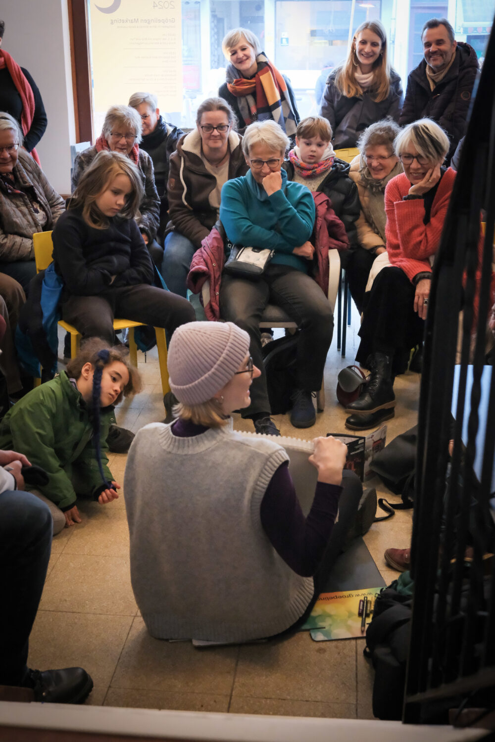 Eine Person sitzt uns mit dem Rücken zugewandt auf dem Boden. Sie zeigt etwas auf einem Zeichenblock. Viele Menschen schauen in ihre Reichtung und sitzen auf Stühlen oder auf dem Fußboden vor ihr.