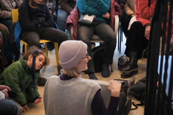 Eine Person sitzt uns mit dem Rücken zugewandt auf dem Boden. Sie zeigt etwas auf einem Zeichenblock. Viele Menschen schauen in ihre Reichtung und sitzen auf Stühlen oder auf dem Fußboden vor ihr.