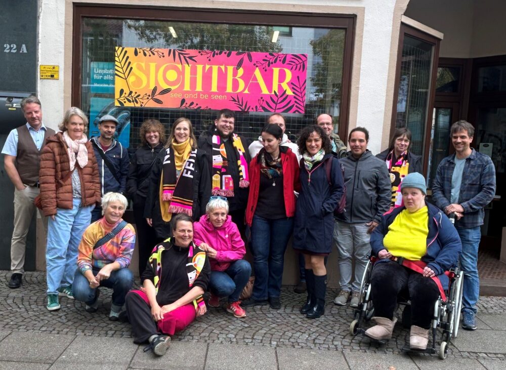 Vor einem Ladenlokal stehen und hocken viele Personen und lächeln für ein Gruppenfoto. Im Fenster des Lokals hängt ein Banner auf dem "Sichtbar" geschrieben steht.