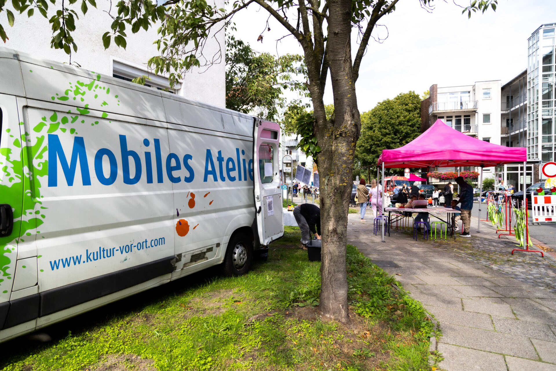 In einer Wohngegend befindet sich ein weißer Bulli mit der Aufschrift "Mobiles Atelier" und daneben ein Baum. Im Hintergrund ist ein pinker Pavillon und sind Menschen zu sehen.