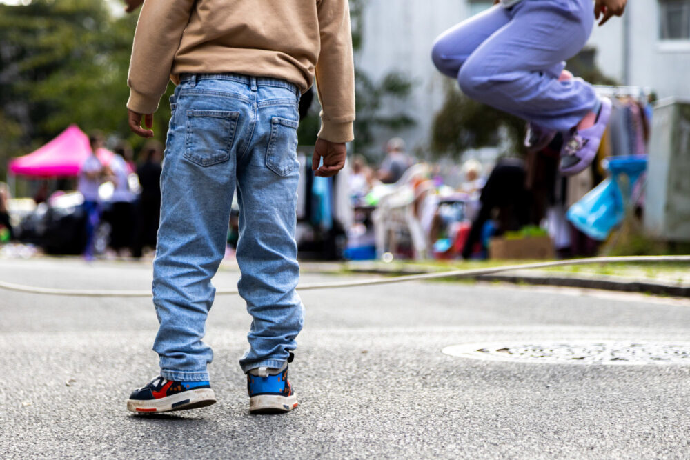 Mitten auf einer Staße sind Kinderbeine zu sehen. Eins der Kinder hüpft in die Luft.