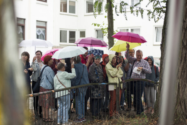 Eine Gruppe Menschen steht dicht gedrängt zusammen unter bunten Regenschirmen, um sich vor dem Regen zu schützen. Sie schauen in verschiedene Richtungen, manche von ihnen lächeln dabei.