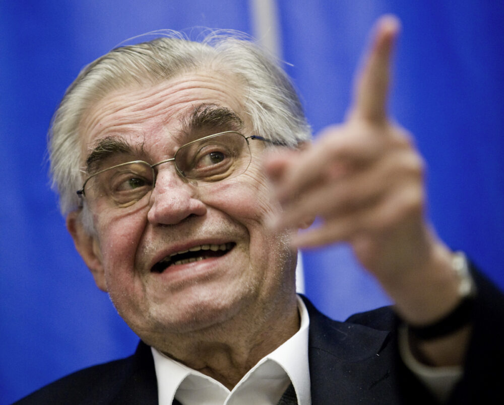 Vor einem blauen Vorhang befindet sich eine grauharige Person mit Brille. Die Person hat einen geöffneten Mund schaut links zur Seite und zeigt mit der linken Hand mit ausgesteckten Zeigefinger nach links.