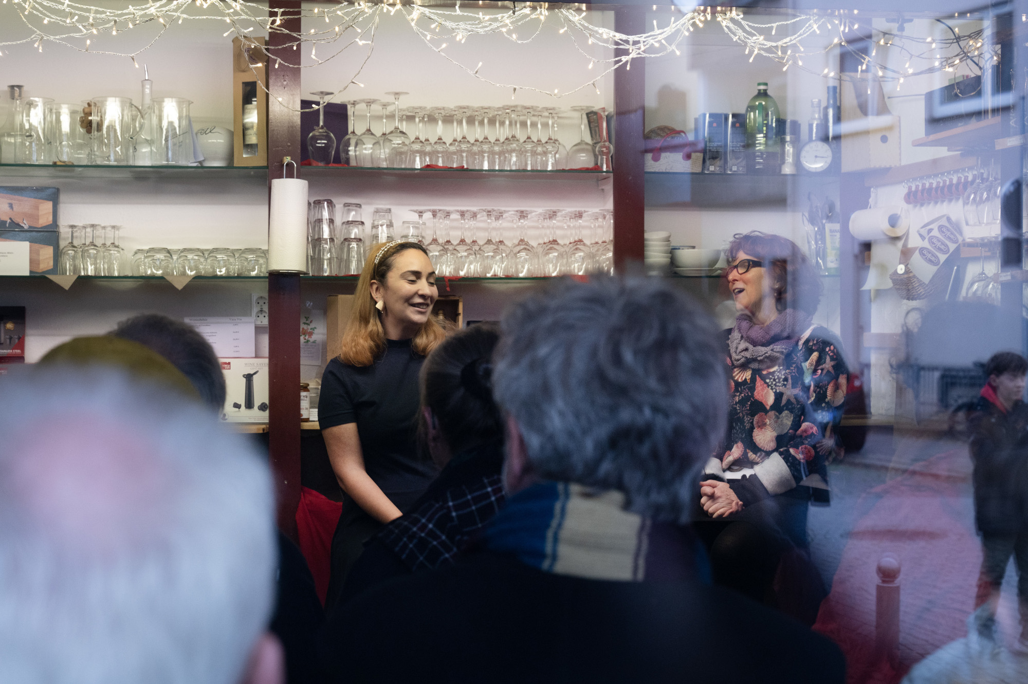 In einer Weinhandlung mit Regalen mit Weingläsern sitzen Menschen, die nur von hinten zu sehen sind. Direkt vor dem Weinregal sitzen, dem Publikum zugewandt zwei Personen, die beide lächeln und sprechen.