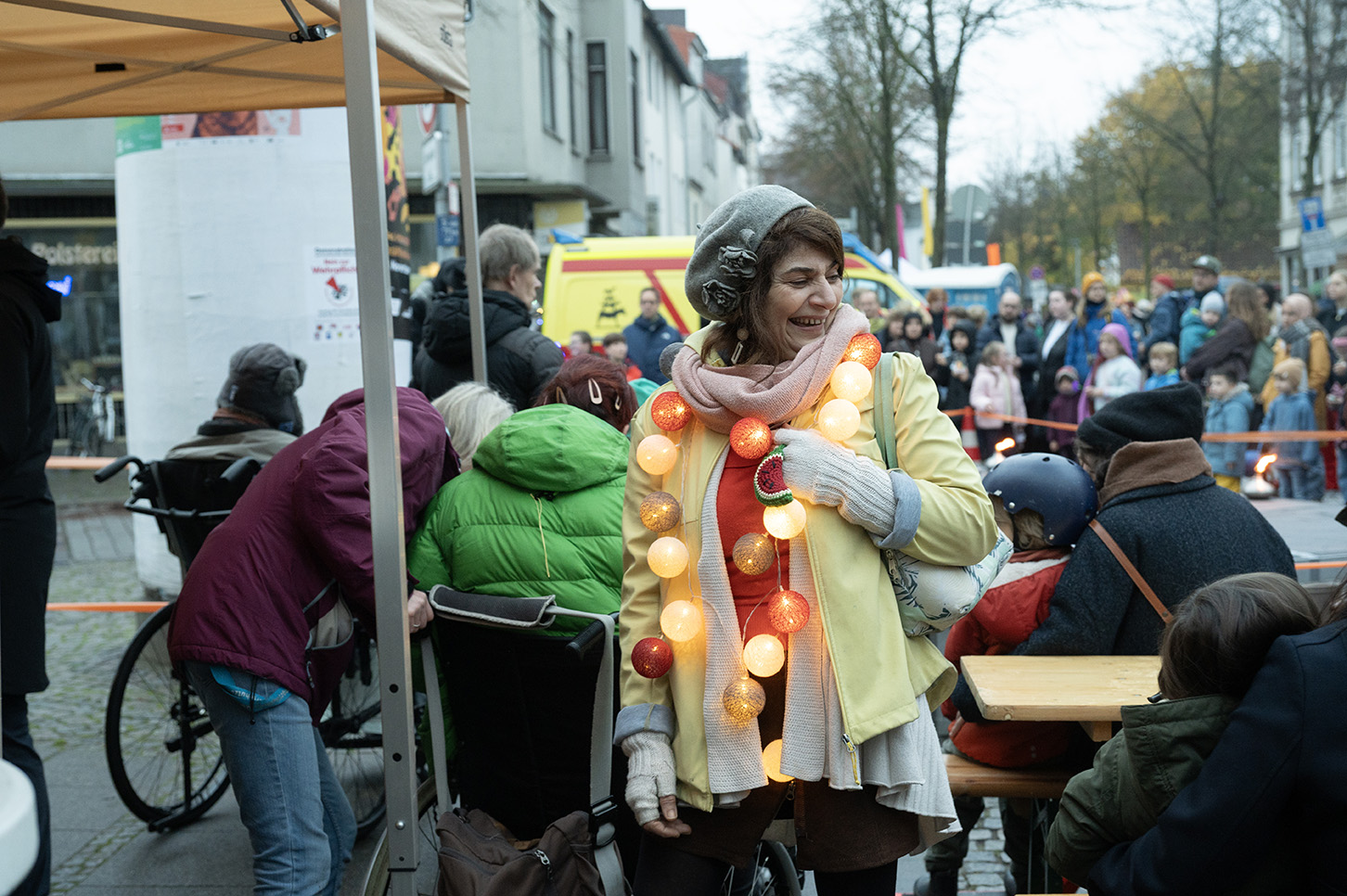 Auf einer belebten Straße sind Menschen an Biertischen im Hintergrund zu sehen. Davor steht eine Person mit Mütze und Schal und Lichterkette und lächelt zur Seite. Links sind ausschnittsweise ein Pavillon und eine Litfasssäule zu sehen.