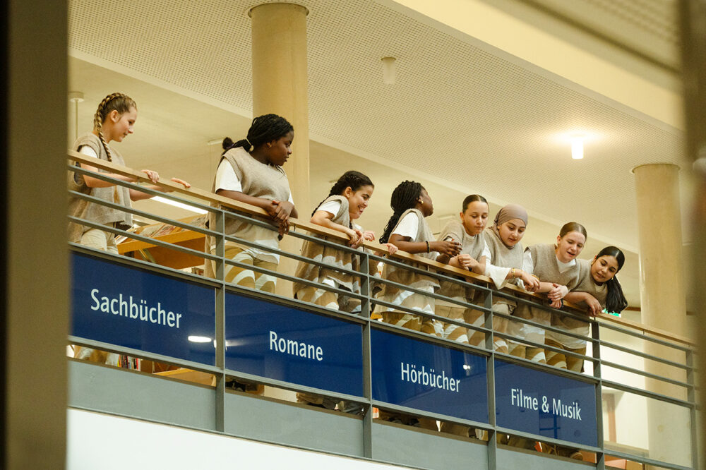 Es ist das Geländer ein einem Gebäude zu sehen. Mehrere Jugendliche lehnen am Geländer und schauen herunter, die meisten in eine Richtung.