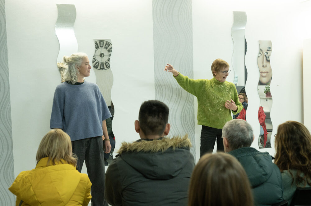 Vor einer hellen Wand mit Spiegeln stehen zwei Personen. Eine schaut die anderen an, die andere schaut in das vor ihnen sitzende Publikum. Sie trägt einen grün-glitzernden Pulli und gestikuliert mit den Armen und spricht.