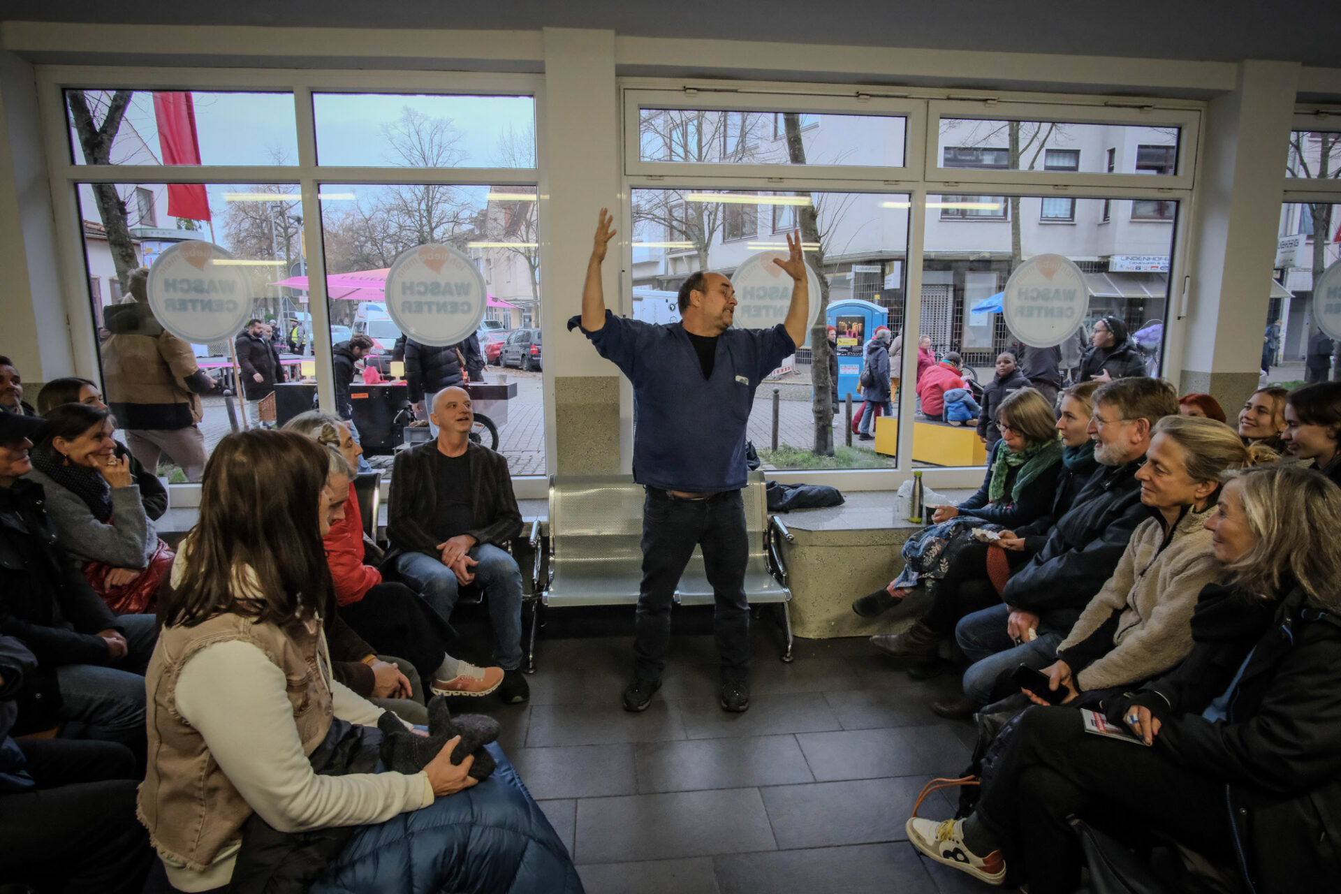 Ein Erzähler hält gestikulierend beide Arme hoch neben seinen Kopf. Links und rechts von ihm sitzt Publikum und hört ihm zu.