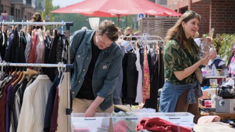 Ein Mann beugt sich über einen Flohmarktstand. Rechts von ihm unterhält sich eine Frau. Im Hintergrund ist der Schirm eines Sonnenschirms zu sehen. Kleidung hängt auf Kleiderbügeln an Garderobenständern.