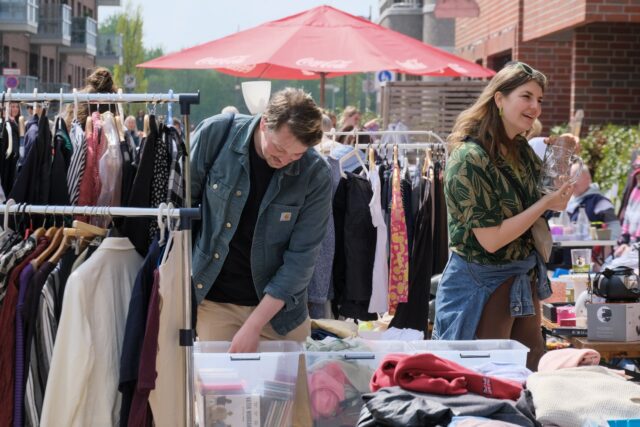 Ein Mann beugt sich über einen Flohmarktstand. Rechts von ihm unterhält sich eine Frau. Im Hintergrund ist der Schirm eines Sonnenschirms zu sehen. Kleidung hängt auf Kleiderbügeln an Garderobenständern.