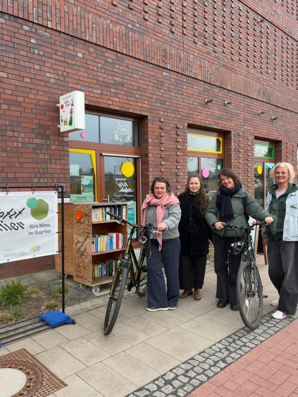Vier Personen stehen vor einem Gebäude mit Fensterfronten. Zwei Personen haben ein Rad dabei. Neben der Eingangstür befindet sich ein Bücherregal auf Rollen. Es ist ein Banner zu sehen, auf dem steht: Jetzt Hier fürs Klima im Quartier.
