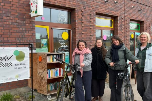 Vier Personen stehen vor einem Gebäude mit Fensterfronten. Zwei Personen haben ein Rad dabei. Neben der Eingangstür befindet sich ein Bücherregal auf Rollen. Es ist ein Banner zu sehen, auf dem steht: Jetzt Hier fürs Klima im Quartier.