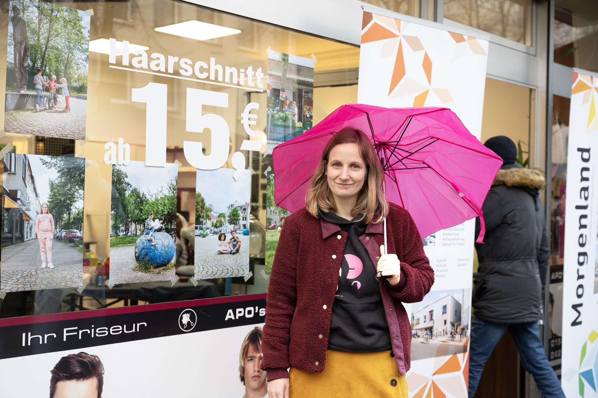Vor der Fensterfront eines Frisörsalons steht eine lächelnde Person, die einen pinken Regenschirm hält.
