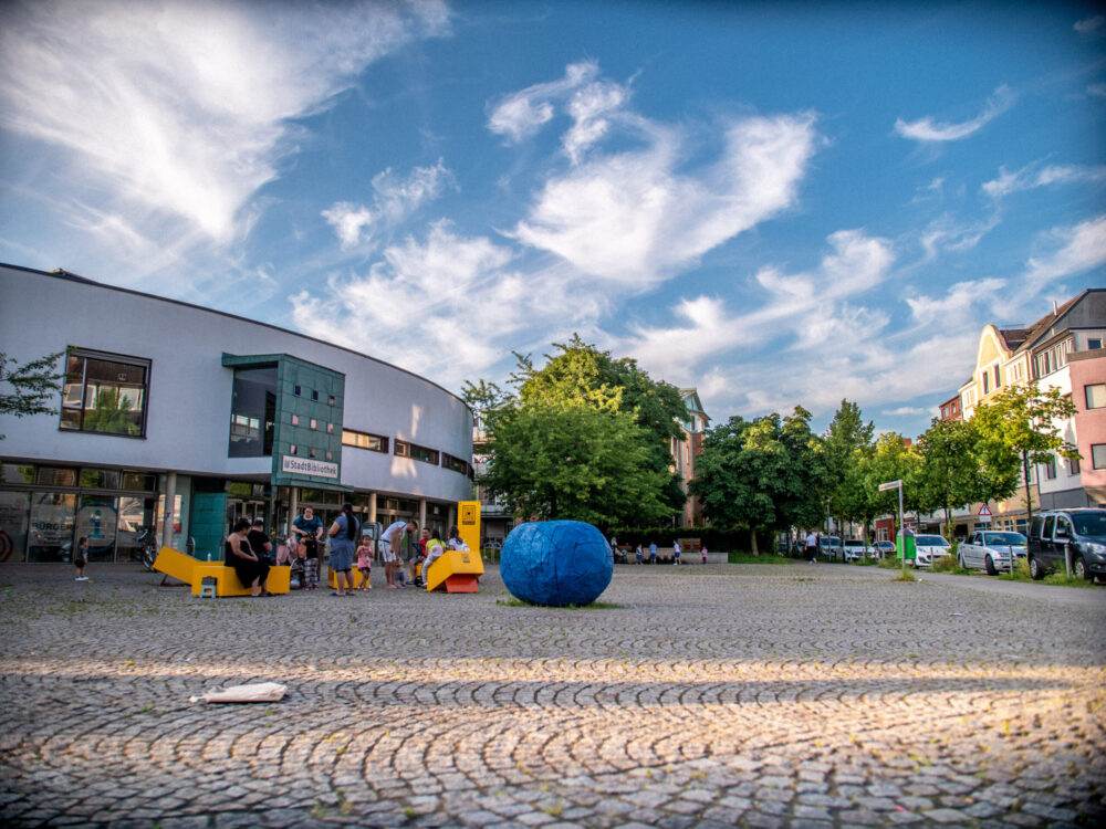 Auf einem großen gepflasterten Platz befindet sich eine runde blaue Skulptur. Daneben stehen und sitzen Personen auf gelben Stadtmöbeln. Links davon befindet sich das ovale Gebäude einer Bibliothek. Im Hintergrund sind Häuser und Bäume zu sehen.