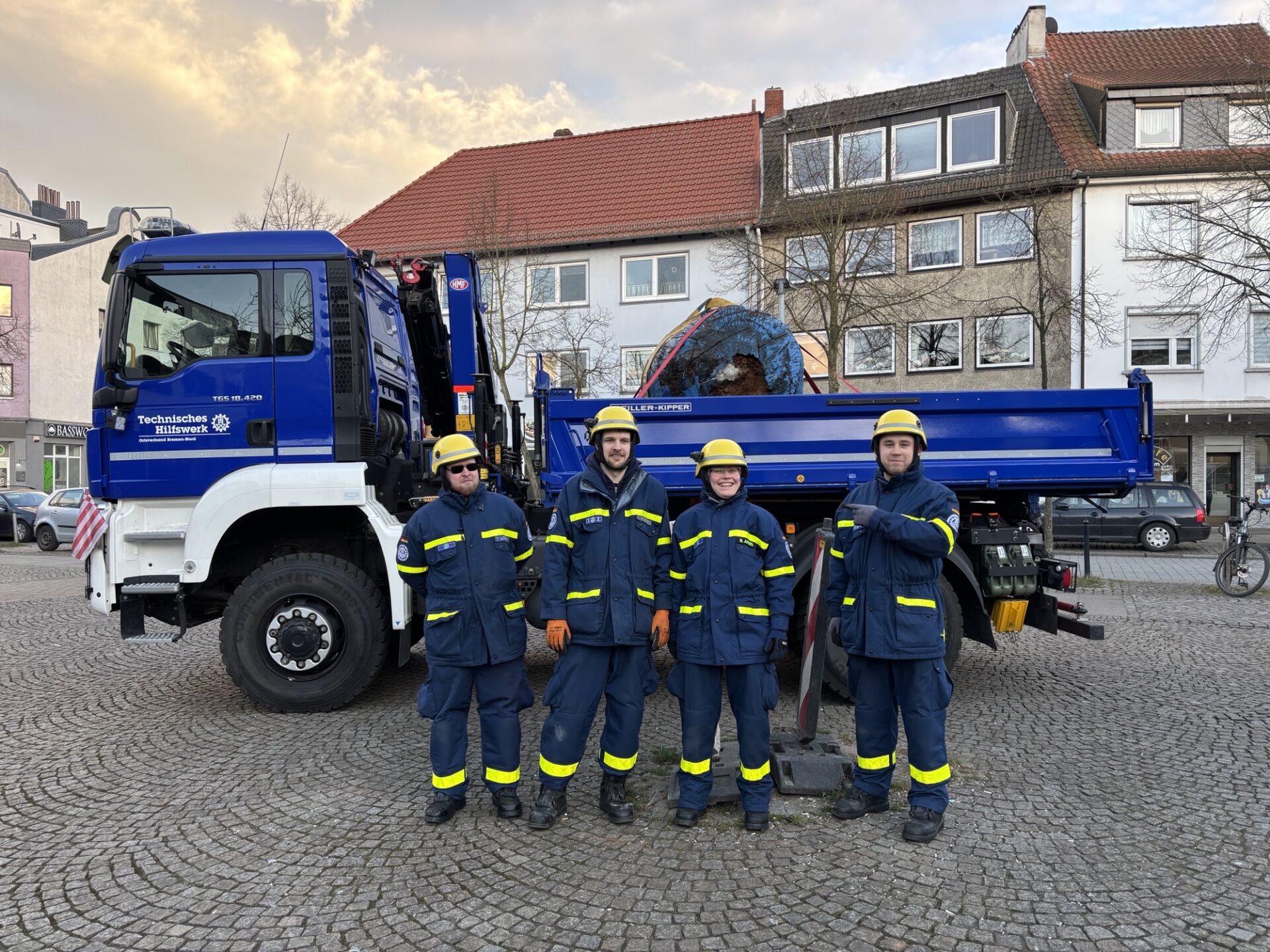 Fünf Mitarbeiter des Technischen Hilfswerks (THW) stehen vor einem THW-Fahrzeug. Sie tragen Schutzkleidung mit Reflexstreifen und Schutzhelme. Im Hintergrund ist das Fahrzeug mit einem Kran und einer blauen Kugel auf dem Laderaum zu sehen. Es ist der blaue Planet vom Gröpelinger Bibliotheksplatz.