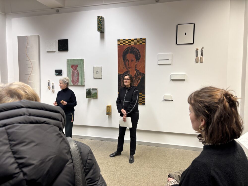 Zwei Personen stehen vor einer Wand, an der Kunstwerke hängen. Eine der Personen spricht, die zweite hält Papiere in der Hand. Zwei weitere Personen sind von hinten zu sehen. sie hören zu.