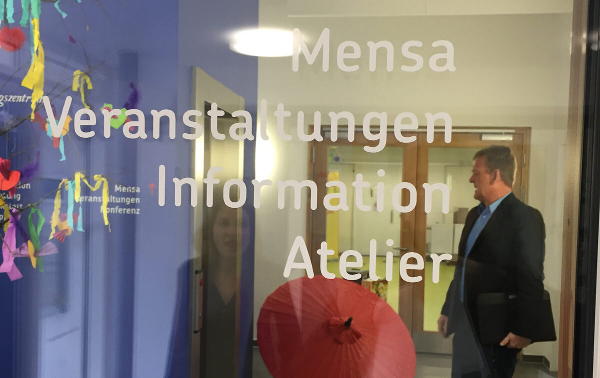 Durch eine Glasscheiben ist ein bunter Hausflur zu sehen und eine Person, die sich etwas an einer Wand anschaut. Die Glasscheibe ist voller Buchstaben.