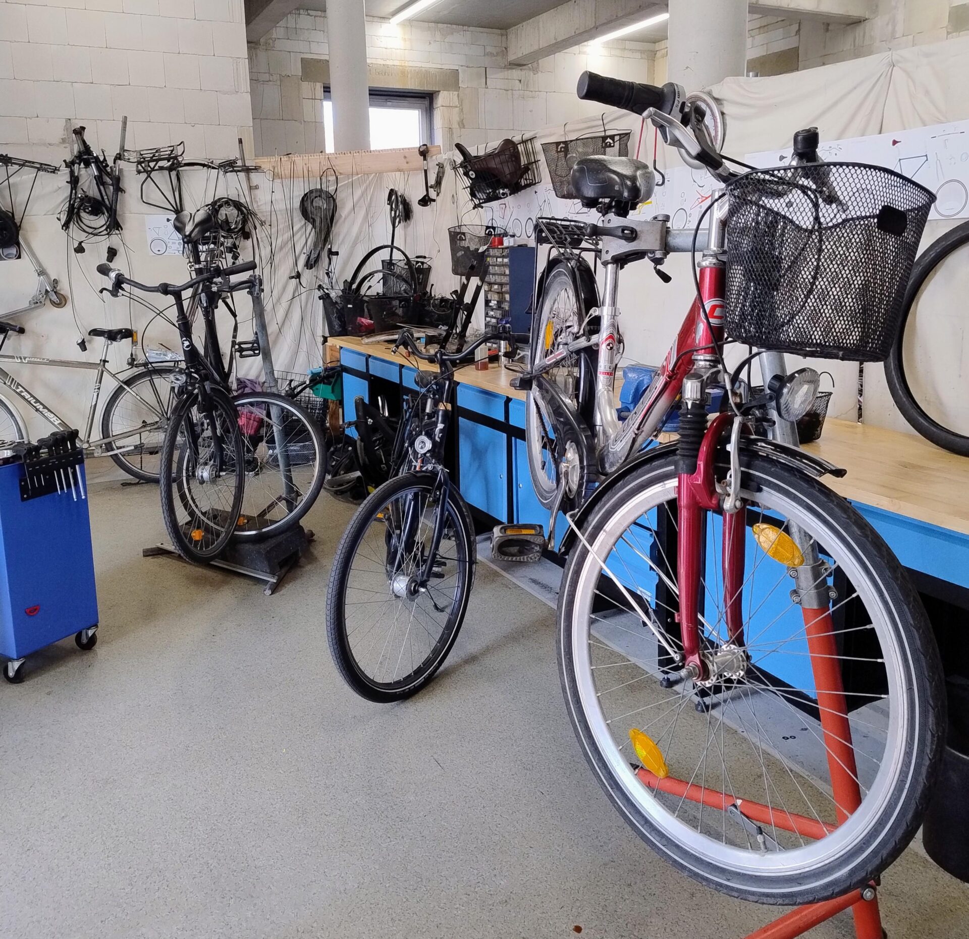 In einer Fahrradwerkstatt stehen mehrere Fahrräder auf dem Boden. Eins der Räder ist in einer Halterung befestigt. Blaue Werkzeugschränke sind im Raum verteilt.
