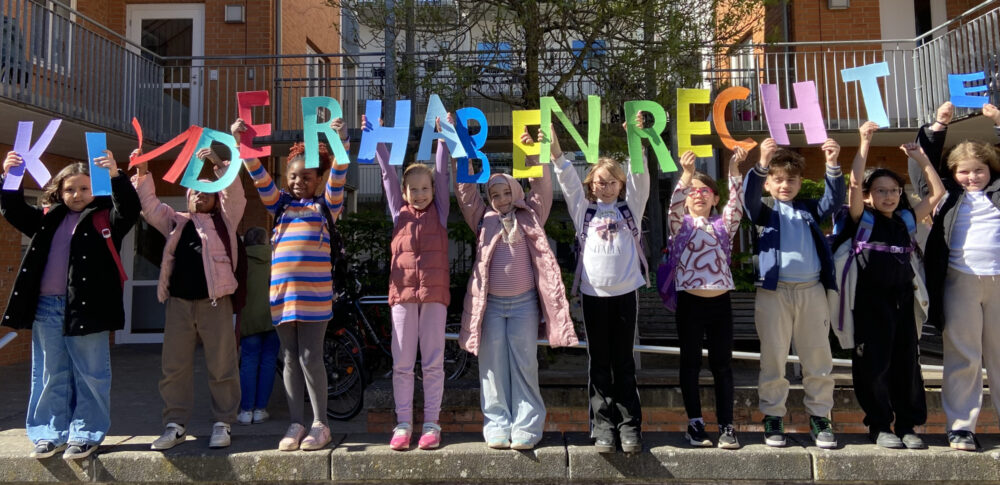 Auf einer Mauer stehen viele Kinder. Jedes Kind hält einen Buchstaben aus angemalten Karton hoch. Die Buchstaben ergeben den Satz "Kinder haben Rechte"