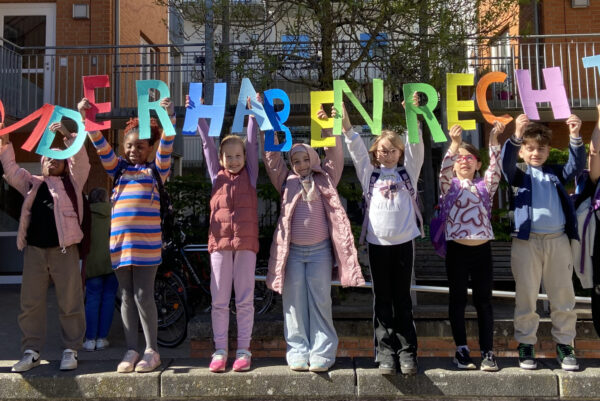 Auf einer Mauer stehen viele Kinder. Jedes Kind hält einen Buchstaben aus angemalten Karton hoch. Die Buchstaben ergeben den Satz "Kinder haben Rechte"