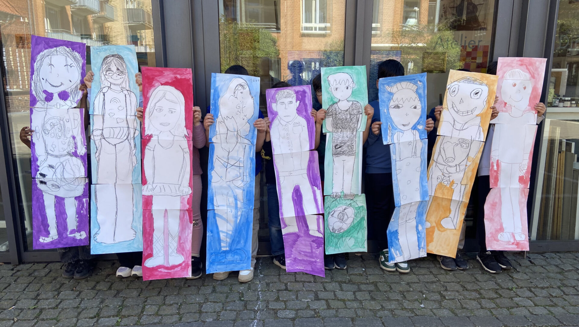 Neun Kinder halten vor dem eigenen Körper lebensgroße Bilder auf Karton auf dem sie selber als Zeichnung zu sehen sind.