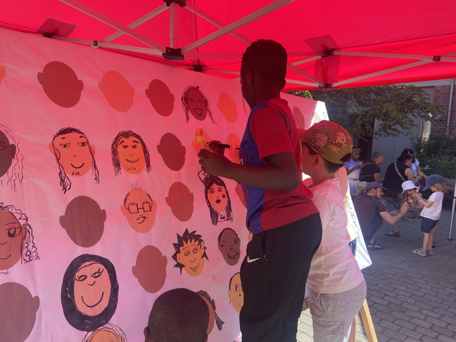 Kinder stehen unter einem Pavillon und Zeichnen Gesichter auf einer Plakatwand. Im hintergrund sind noch mehr Kinde und auch Erwachsene zu sehen.